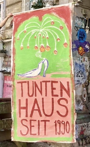 Gemaltes Transparent vor einem unsanierten Haus. Darauf ein Kronenleuchter, ein Stöckelschuh und der Text "Tuntenhaus seit 1990"