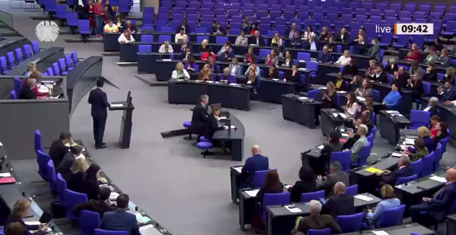Zu sehen ist der Plenarsaal des Bundestages, der FDP-Generalsekretär Djir-sarai am Redepult. Zwischenfrage von Gesine Lötzsch, Linke, aus einer der hinteren Reihen im Plenum. Vor ihr sitzen einige gelangweilt in Smartphones guckende MdB der SPD, um sie herum einige linke MdB.