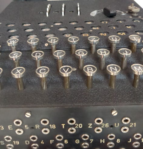 Tastatur der Enigma: runde Tasten mit Buchstaben an einem schwarzen Gehäuse, dahinter verschiedene Schalter, vorn mit Buchstaben und Zahlen beschrifteten runde Löcher.