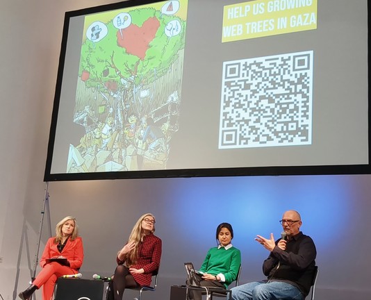 Vier Menschen auf einem.Podium, eine Person spricht. Über ihnen eine Leinwand  mit einem gezeichneten Bild eines Baums, ein QR Code und Text „Help us growing web trees in Gaza“