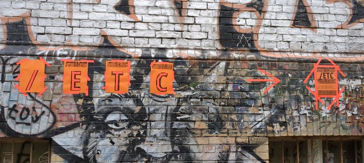 Brick wall with grey and brown graffiti. On it four orange sheets of paper in a row, each with one letter: /etc.
To the right an orange arrow pointing right to another printout on orange paper, to small to read.