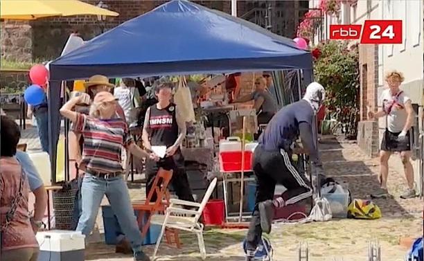 Eine Person in Jeans und gestreiftem T-Shirt hält in der erhobenen rechten Hand einen Kochtopf und richtet sich in Richtung einer dunkel gekleideten, laufenden Person, die von hinten zu sehen ist. Mehrere Personen sehen zu, darunter eine mit einem T-Shirt, auf dem in Großbuchstaben FMNSM steht. Im Hintergrund Menschen, Luftballons, die Sonne scheint, es sind Stühle und ein Zeltdach zu sehen. Ganz hinten und an der Seite sind Gebäude zu erkennen.