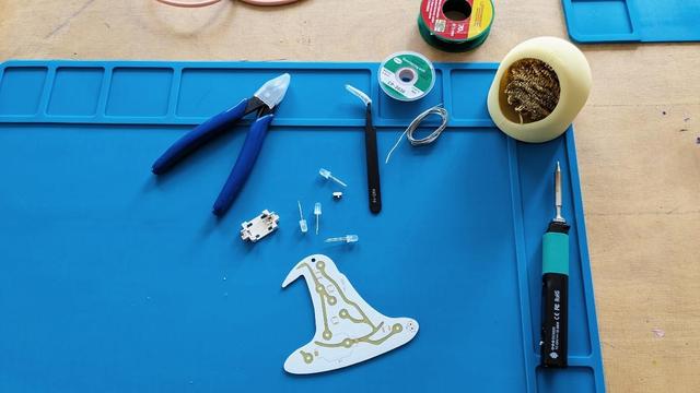 Arbeitsplatz zum Löten mit Lötzinn, Zangen, einer kleinen Platine in Form des Haecksen-Logos, dem Haecksenhut und diversen dazu nötigen Kleinteilen.