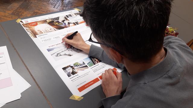 Blick über die Schulter eines Mannes. Auf den Ausdruck der Startseite der Deutschen Bahn schreibt er mit einem Filzstift das Wort "Bildergalerie" über ein Foto.