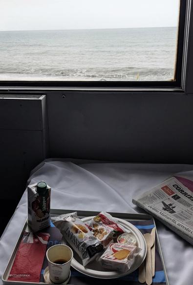 Fußende eines mit einem weißen Laken bezogenen Betts in einem Zugabteil. Darüber das Zugfenster mit Blick.aufs Meer. Auf dem Bett ein Tablett mit Frühstück und Zeitung.