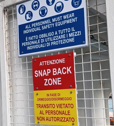 Warnschilder an einem weißen Gitter
'All personnel must wear individual safety equipment'
'Attenzione Snap Back Zone'
'In fase di ormeggio/disormeggio transito vietato al personale non autorizzato'