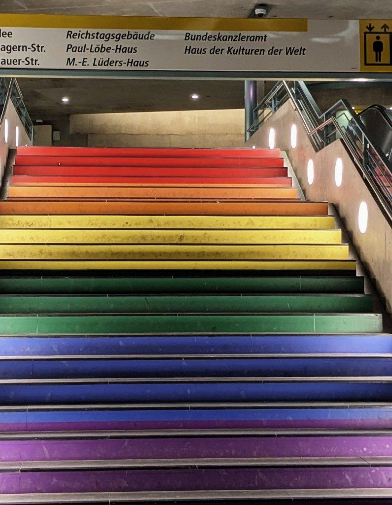 Treppe von unten fotografiert. Die Stufen sind von vorn in Regenbogenfarben eingefärbt. Rechts ist eine Rolltreppe, oben ein Schild der BVG mit Hinweisen zum Reichstagsgebäude, Paul-Löbe-Haus, Bundeskanzleramt, Haus der Kulturen der Welt u.a.