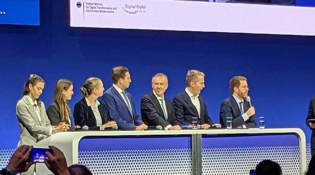 Seven people behind a table on stage. Three women on the left, four men on the right, taking up at least two thirds of the space.