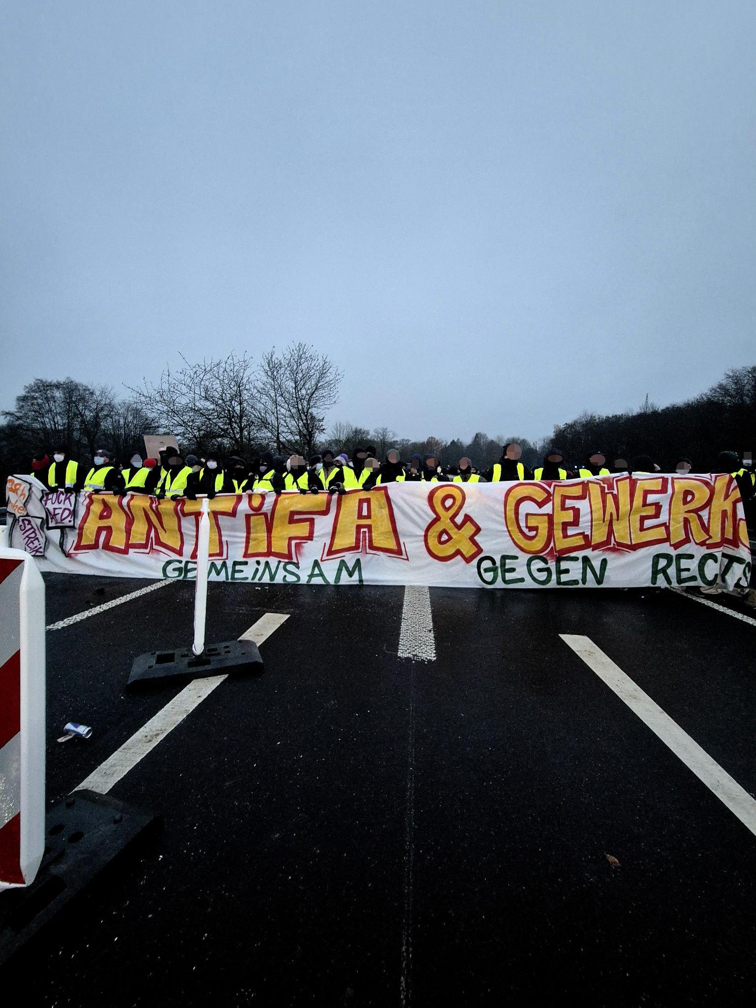 Eine größere Gruppe Menschen in gelben Warnwesten blockiert eine Straße und hält ein großes Banner. Darauf steht in roten und gelben Buchstaben „ANTIFA & GEWERKSCHAFTEN“, darunter in Grün „GEMEINSAM GEGEN RECHTS“. Die Umgebung wirkt winterlich und grau, Bäume ohne Laub stehen im Hintergrund.