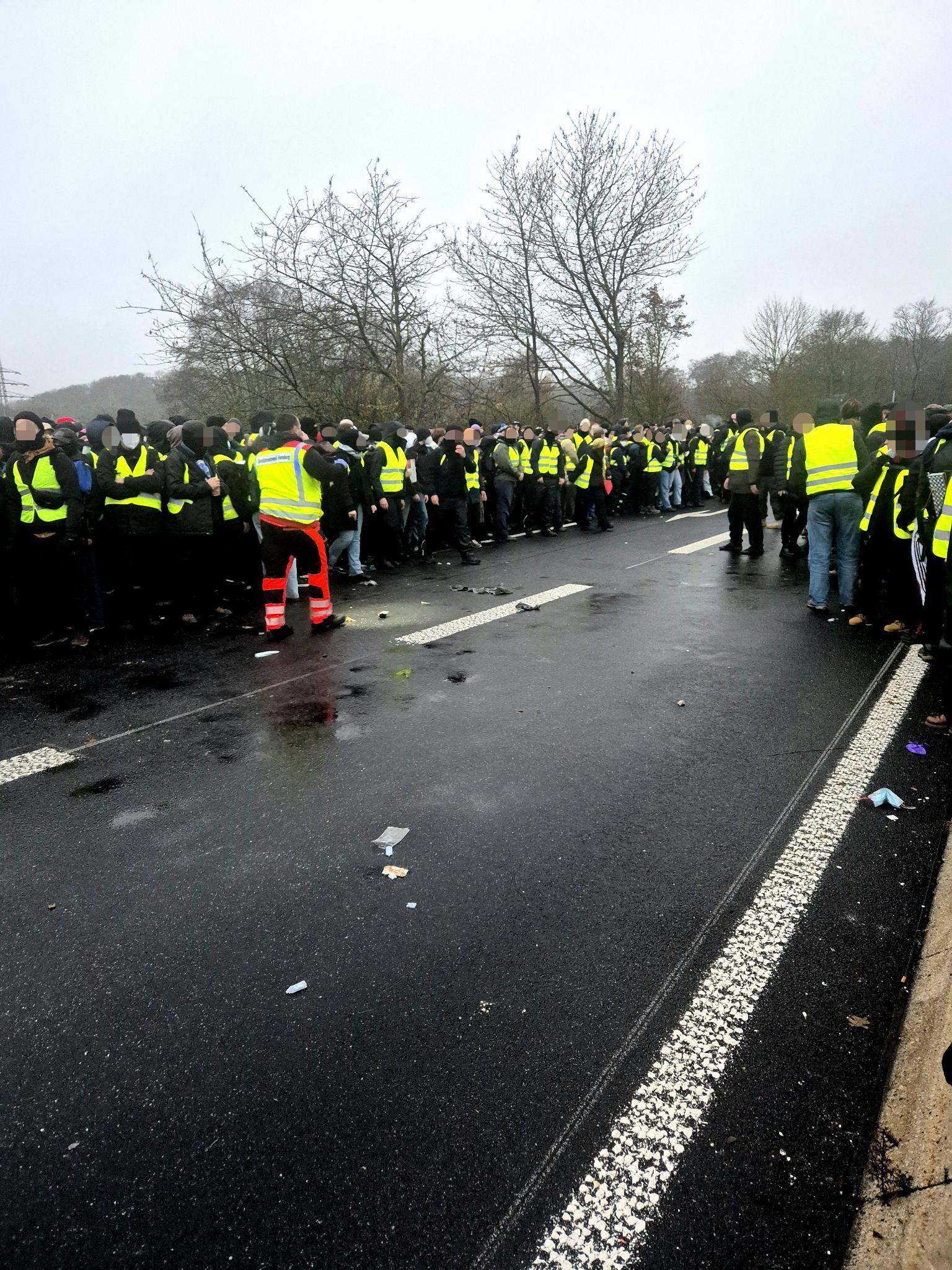 Eine große Gruppe von Menschen in gelben Warnwesten blockiert eine nasse Straße. Auf dem Bild bilden sie eine Rettungsgasse.