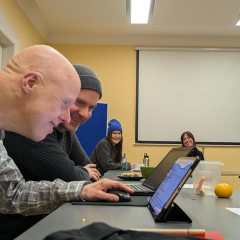 Zwei Männer schauen lachend auf ein Tablet, beobachtet von zwei Frauen, die ebenfalls lachen.