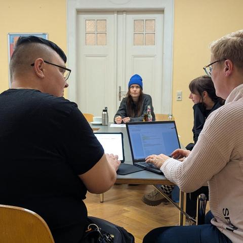 Vor einem Tablet und einem Laptop sitzen zwei Frauen. Eine der beiden tippt. Ihnen gegenüber sitzen zwei weitere Frauen. Sie schauen ihnen dabei zu.