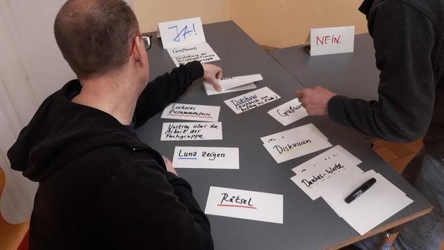 Zwei Personen sortieren Zettel an einem Tisch, auf denen Wörter wie Luna zeigen, Diashow, Dankesworte, Rätsel oder lockeres Zusammensein stehen.