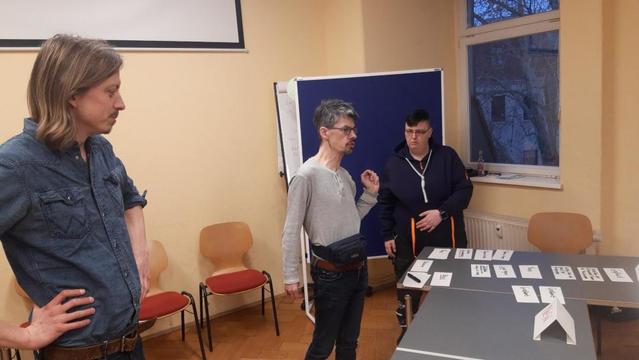 Ein Fachgruppen-Mitglied spricht in einem Seminarraum. Zwei Personen hören zu.