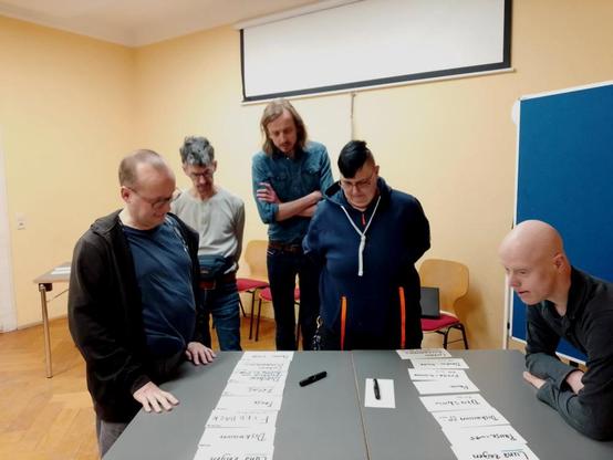 Fünf Personen stehen in einem Seminarraum um einen Tisch und begutachten Zettel, die in zwei langen Reihen vor ihnen liegen.