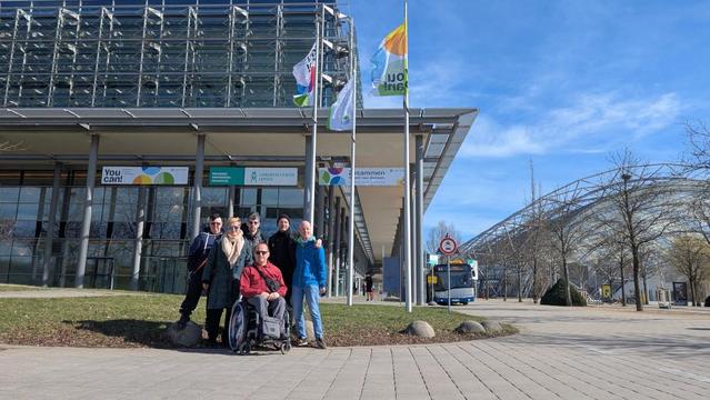 Die sechs Personen aus einer anderen Perspektive. Im Hintergrund sieht man jetzt Fahnenmasten und einige Banner. Auf einem steht zum Beispiel "You can!" Auf einem anderen "Zusammen Arbeit neu denken" Etwas weiter im Hintergrund die markante Glaskuppel der Messe Leipzig.