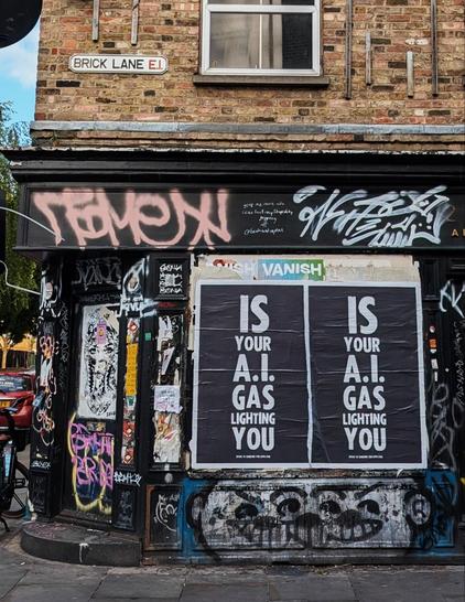 Unterer Teil eines mehrgeschossigen gemauerten Hauses an einer Straßenecke. Das Erdgeschoss ist schwarz gestrichen, darauf viel Graffiti. Auf zwei nebeneinander angebrachten Plakaten steht in weißer Schrift auf schwarzem Hintergrund 'Is your A.I. gaslighting you'
Weiter oben  am Haus ist ein Straßenschild zu sehen, auf dem Brick Lane steht.
