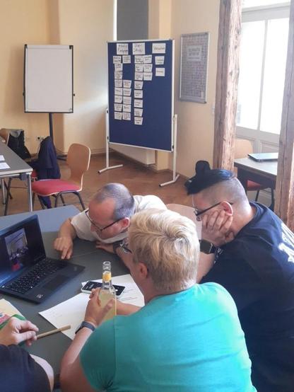 In einem Seminarraum sitzen vier Menschen um einen Laptop und schauen auf den Bildschirm, auf dem ein Foto zu erkennen ist. Im Hintergrund steht eine Pinnwand mit vielen Karteikarten und ein Flipchart.