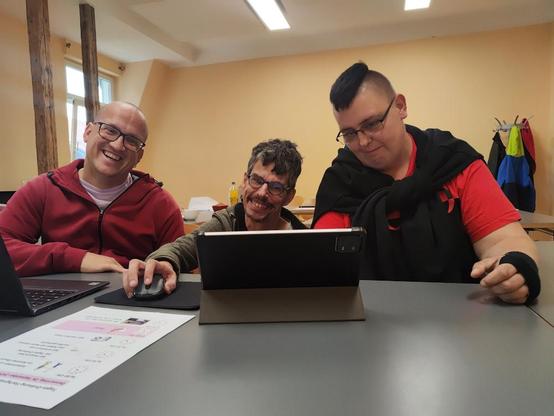In einem Seminarraum sitzen drei gut gelaunte Menschen vor einem Laptop und einem Tablet. Vor ihnen liegt auch ein Ausdruck, der aussieht wie eine Tagesordnung.