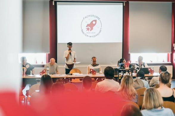 In einem großen Saal steht ein Mann mit einem Mikrofon und spricht zum Publikum. Links und Rechts von ihm sitzen 5 Personen. Im Hintergrund sieht man eine Leinwand, auf die eine große Rakete projiziert ist. Dabei steht: "Fachgruppe - Raketen-Wissenschaft - 2023 - 2026."