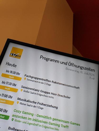 Auf einem Monitor der Villa werden die aktuellen Veranstaltungen angezeigt. Ganz oben steht "Fachgruppentreffen Raketenwissenschaft, Seminarraum 3. Etage." Darunter sieht man Termine wie "Seniorentanz", "Musikalische Früherziehung" oder "Cozy Gaming".